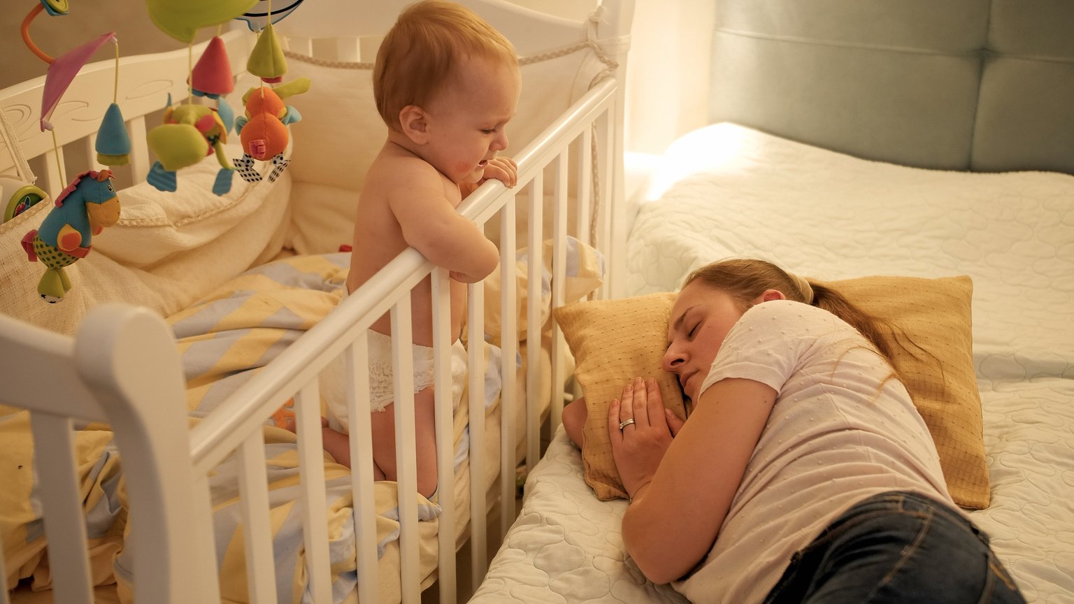 Young Tired Mother Fell Asleep Next to Her Baby Crying in Bed. Concept of Parenting, Parent Fatigue and Children Development.